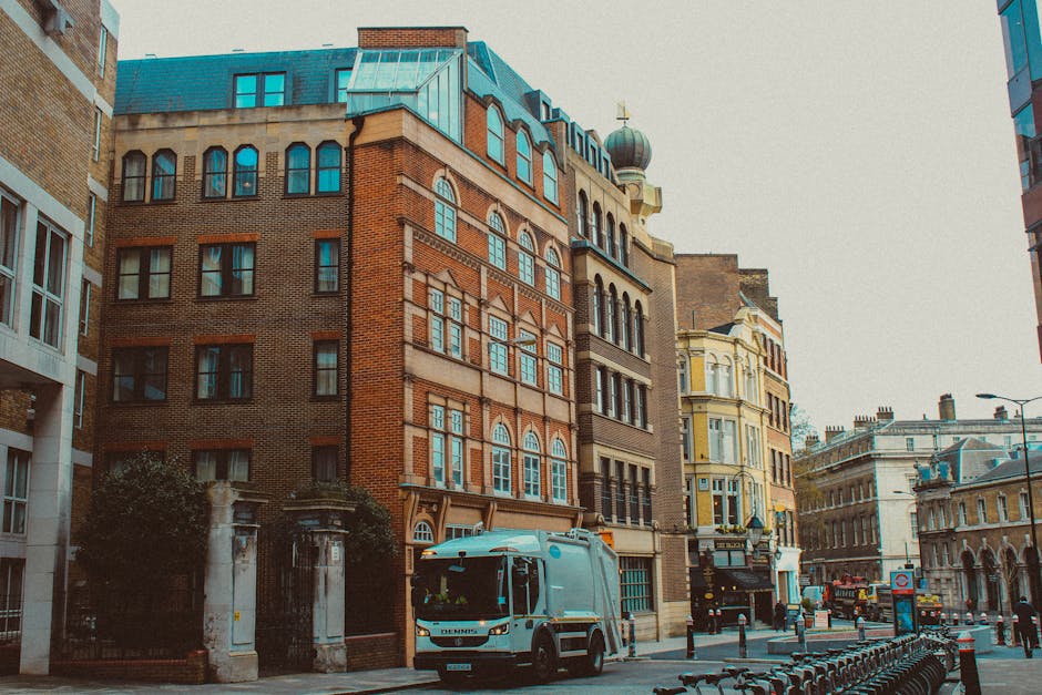 A street scene in an urban area with a row of multi-story buildings showcasing a mix of architectural styles and finishes. The central building features red brickwork with decorative window arches and